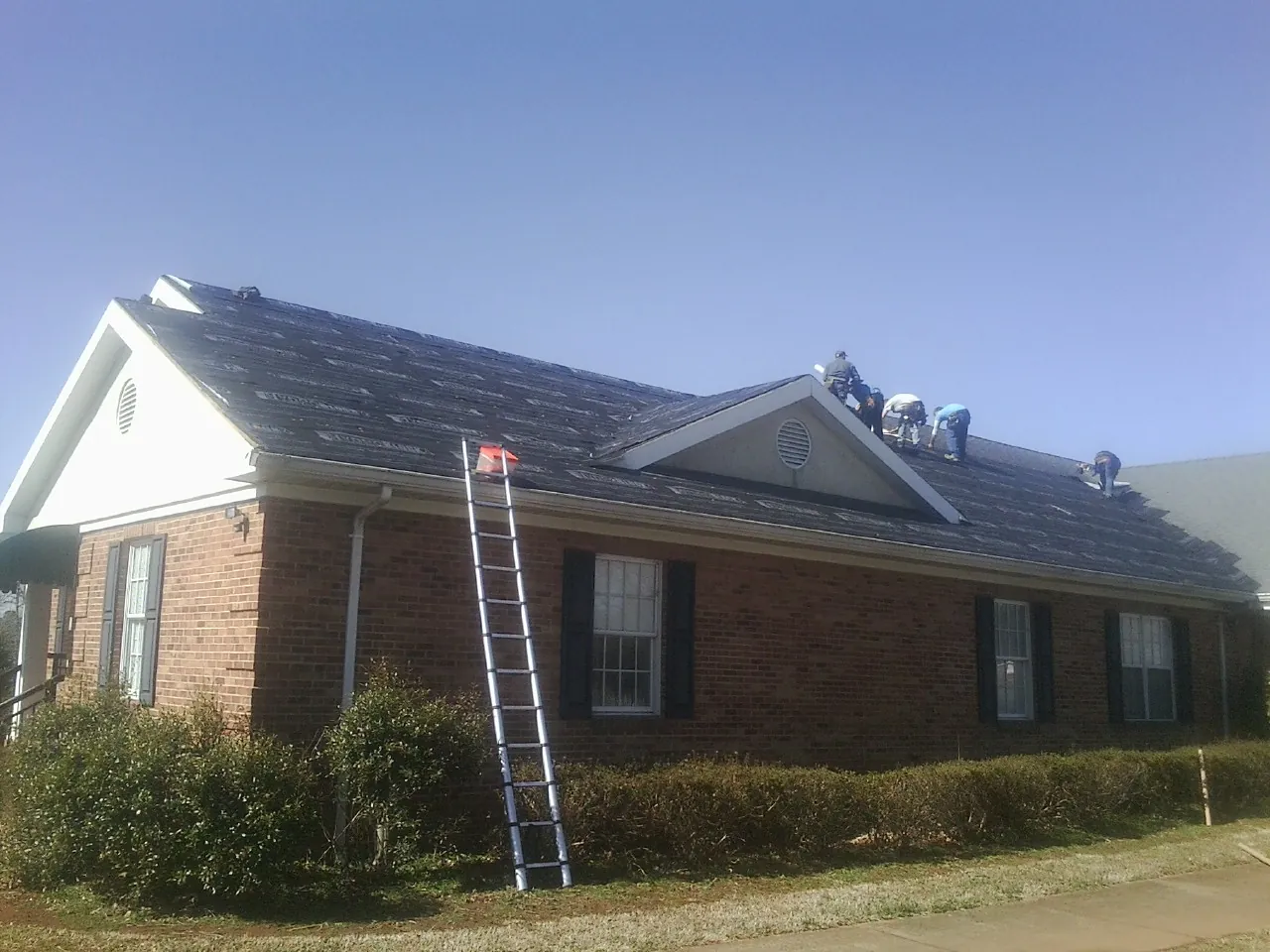 Crew tearing shingles off a brick ranch roof with a ladder set — Metal Roof Repair work by our crew in Hustisford