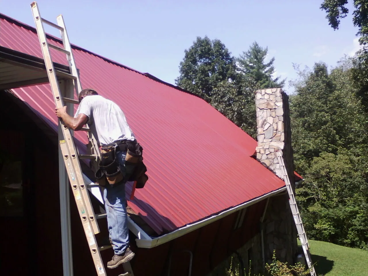 Roofer on a ladder beside a red metal roof and stone chimney — Soffit & Fascia Repair by Roofing Brothers in Hustisford